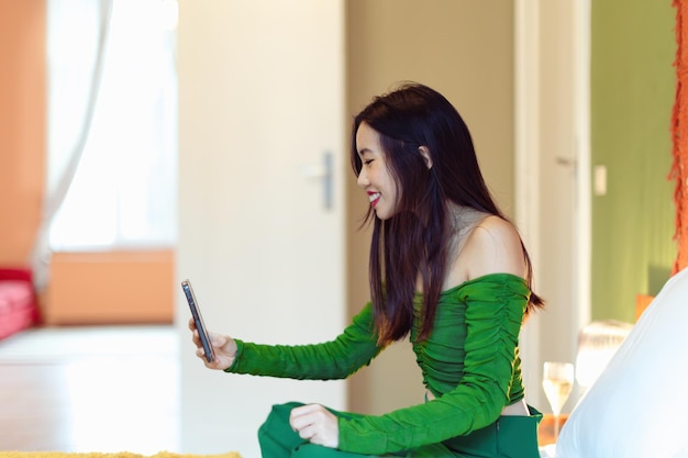Uśmiechnięta azjatycka kobieta siedząca na łóżku w pokoju hotelowym podczas rozmowy wideo z telefonem komórkowym