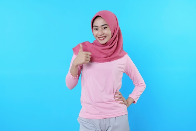 Uśmiechnięta azjatycka kobieta z kciukiem odizolowanym na jasnoniebieskim tle ubrana w hidżab z różową koszulką