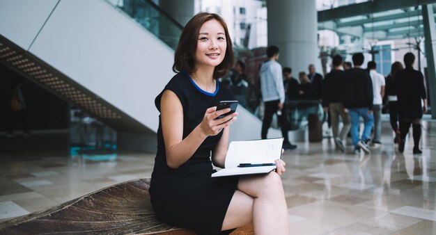 Uśmiechnięta Azjatycka młoda bizneswoman w czarnej eleganckiej sukience z nogami skrzyżowanymi w korytarzu