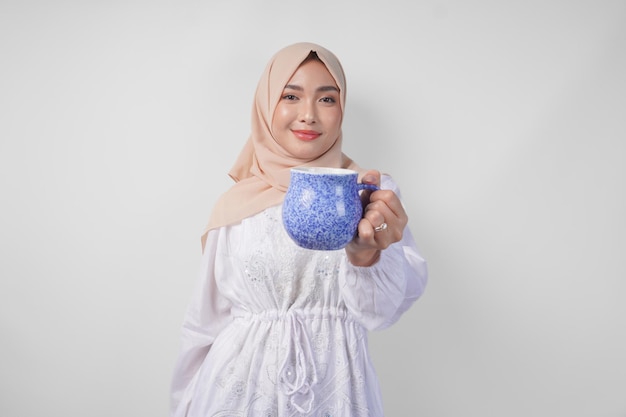 Uśmiechnięta azjatycka muzułmańska kobieta w białej sukience i hidżabie trzymająca i prezentująca wodę lub napój w niebieskim kubku stojąca nad izolowanym białym tłem