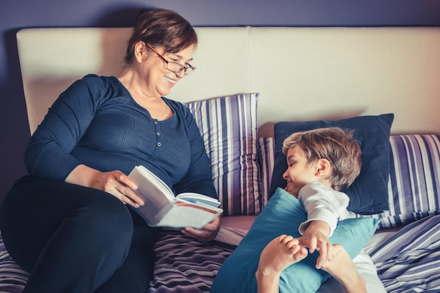 Uśmiechnięta babcia czyta książkę wnukowi na łóżku.