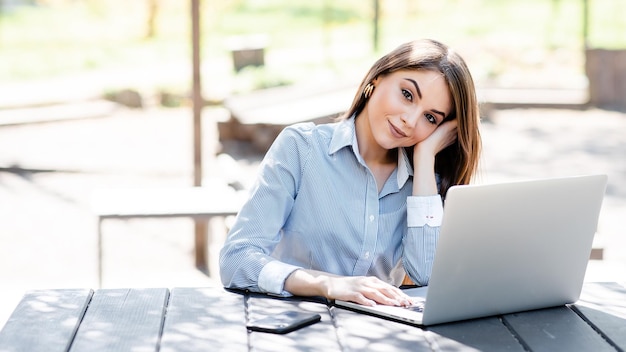 Uśmiechnięta biała bizneswoman pracuje na laptopie na świeżym powietrzu Piękna młoda dziewczyna siedzi przy stole w słonecznym parku i patrzy na kamerę Współczesny styl życia kobiety Koncepcja pracy zdalnej i niezależnej