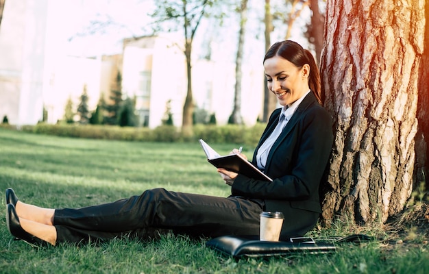 Uśmiechnięta biznesowa kobieta w garniturze z czymś zapisuje w papierowym notesie siedząca podczas przerwy pod drzewem w miejskim parku