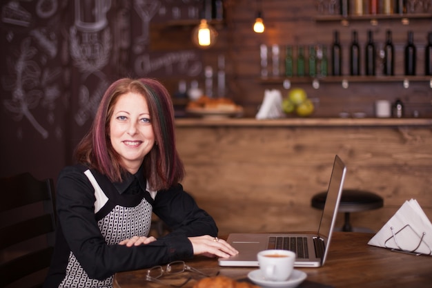 Uśmiechnięta bizneswoman z laptopem w czcionce siedzącej i delektującej się kawą w kawiarni lub restauracji w stylu vintage