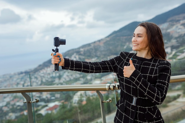 Uśmiechnięta blogerka robi selfie swoim mini aparatem, pokazując idealny gest na tle miasta