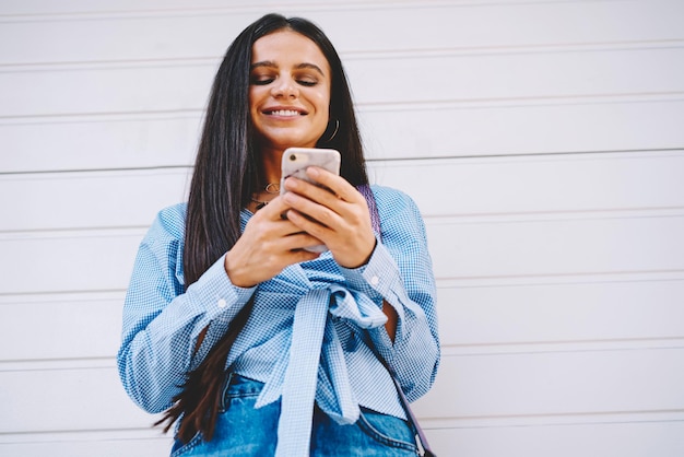 Uśmiechnięta blogerka z długimi włosami, która używa smartfona do czatowania z obserwatorami w sieci społecznościowej