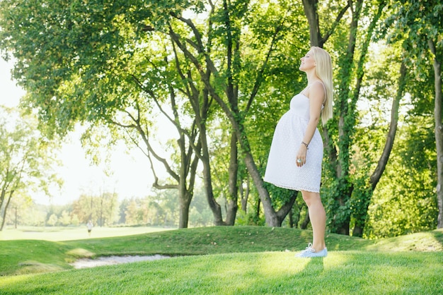 Uśmiechnięta blond kobieta w ciąży w białej sukni wdychać świeże powietrze w parku. Spacer na świeżym powietrzu