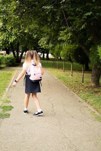 Uśmiechnięta blond uczennica w szkolnym mundurku z różowym plecakiem idzie do szkoły na świeżym powietrzu