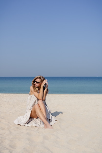 Uśmiechnięta blondynka siedzi na plaży w letni poranek