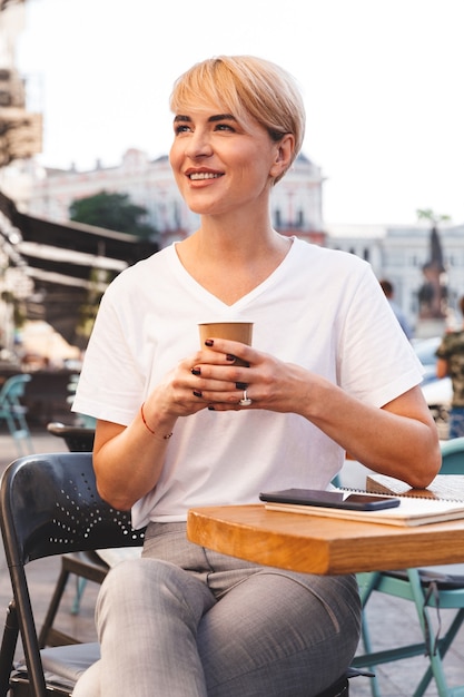 uśmiechnięta blondynka ubrana w zwykłe ubranie siedzi w kawiarni ulicznej latem i pije kawę z papierowego kubka