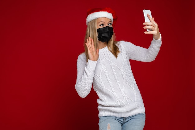 uśmiechnięta blondynka w kapeluszu Santa i czarnej masce ochronnej macha ktoś dzwoni przez telefon komórkowy podczas pandemii. Izoluj na czerwono. Skopiuj miejsce.