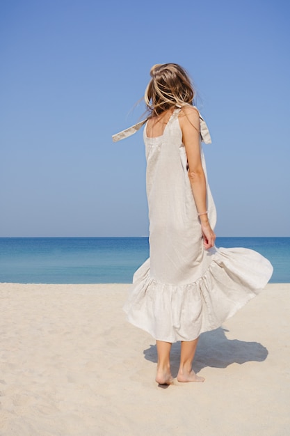 Uśmiechnięta blondynka w lnianej sukience maxi z fruwającymi włosami, skacząca i tańcząca na piaszczystej plaży