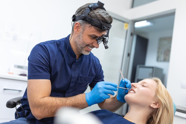Uśmiechnięta brunetka badana przez dentystę w klinice dentystycznej Ręce lekarza trzymającego instrumenty dentystyczne w pobliżu ust pacjenta Zdrowe zęby i koncepcja medycyny