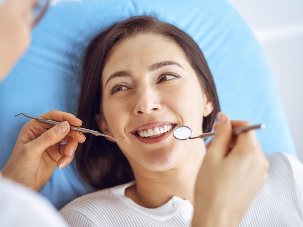 Uśmiechnięta brunetka kobieta badana przez dentystę w klinice dentystycznej. Ręce lekarza trzymającego instrumenty dentystyczne w pobliżu ust pacjenta. Zdrowe zęby i koncepcja medycyny.