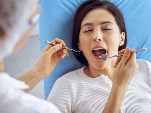 Uśmiechnięta brunetka kobieta badana przez dentystę w klinice dentystycznej. Ręce lekarza trzymającego instrumenty dentystyczne w pobliżu ust pacjenta. Zdrowe zęby i koncepcja medycyny.