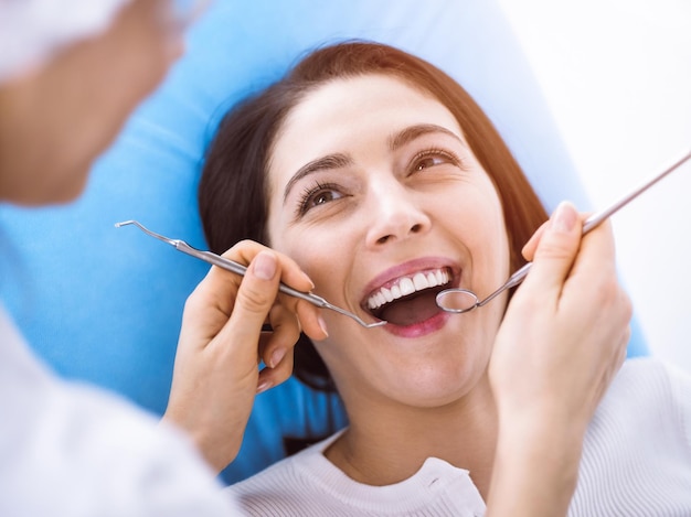Uśmiechnięta brunetka kobieta badana przez dentystę w słonecznej klinice stomatologicznej. Ręce lekarza trzymającego instrumenty dentystyczne w pobliżu ust pacjenta. Zdrowe zęby i koncepcja medycyny.