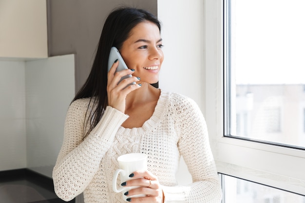 Uśmiechnięta brunetka stoi przy oknie w pomieszczeniu, trzymając filiżankę kawy, rozmawiając przez telefon komórkowy