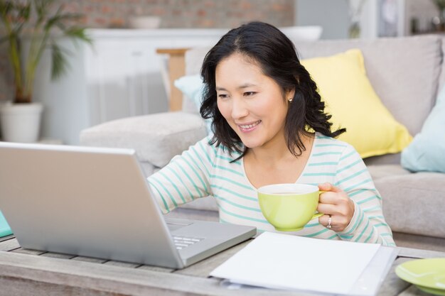 Uśmiechnięta brunetka używa laptop i trzymający kubek w żywym pokoju
