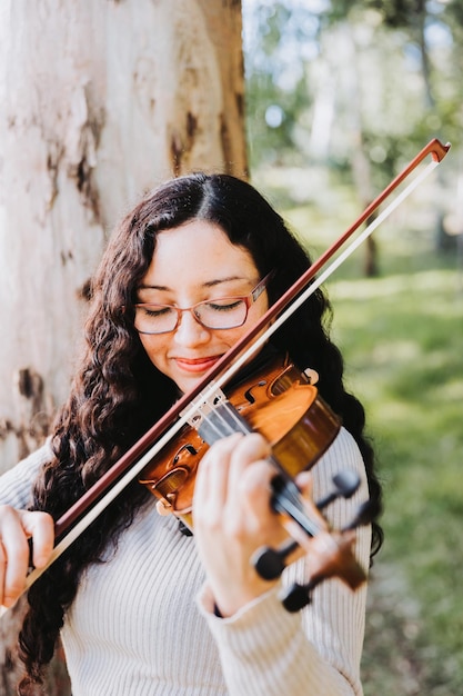 Uśmiechnięta brunetka w okularach gra na skrzypcach na zewnątrz w lesie Vertical