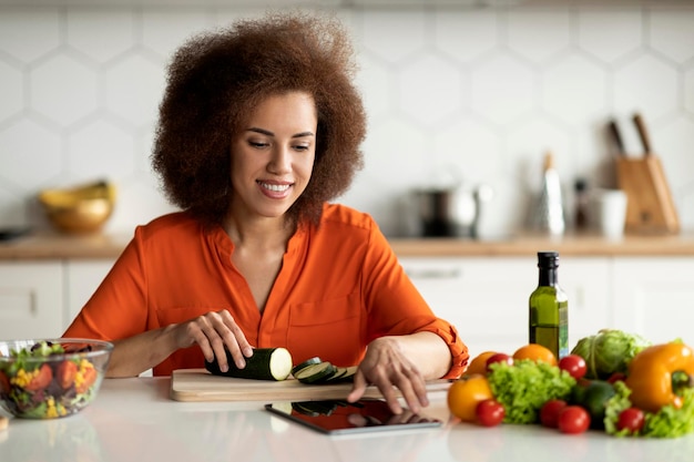 Uśmiechnięta czarna dama sprawdza przepis na cyfrowym tabletie podczas gotowania w kuchni