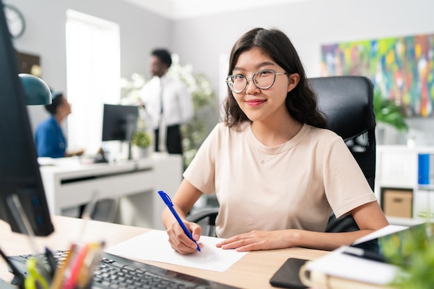 Uśmiechnięta dziewczyna o azjatyckiej koreańskiej urodzie ubrana w luźną koszulkę i okulary siedzi przy biurku