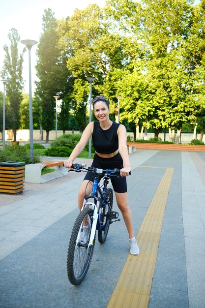 Zdjęcie uśmiechnięta dziewczyna w czarnym topie i szortach prowadząca rower w środowisku miejskim