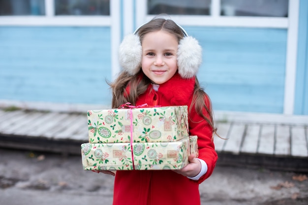 Zdjęcie uśmiechnięta dziewczyna w futrzanych słuchawkach i czerwonym płaszczu stojąca na świątecznej ulicy z prezentami