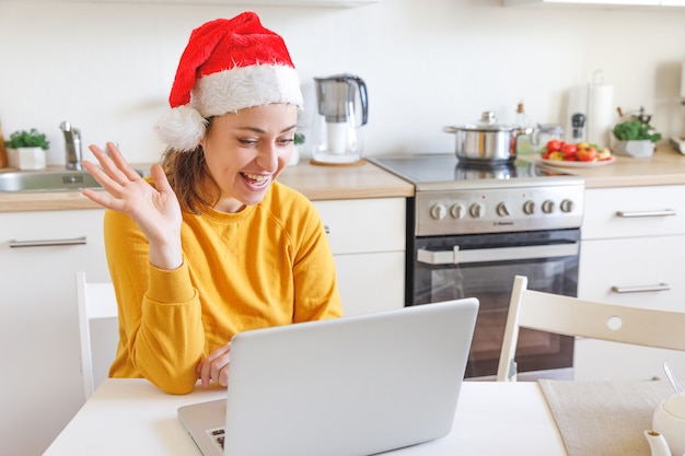 Uśmiechnięta dziewczyna w kapeluszu Santa macha ręką, rozmawiając wideo z rodziną przez kamerę internetową