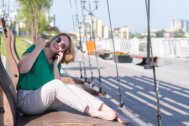 Uśmiechnięta dziewczyna w okularach przeciwsłonecznych rozmawia przez telefon z przyjaciółmi umawiając się na randkę w parku latem
