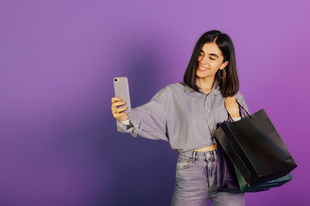 Uśmiechnięta dziewczyna w ubranie, trzymając pakiety torby z zakupami po zakupach i robi selfie na telefonie na białym tle na fioletowej powierzchni