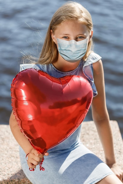 Uśmiechnięta dziewczyna z balonem w kształcie serca w masce medycznej na twarzy