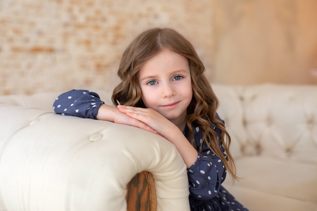 Uśmiechnięta dziewczynka siedzi na kanapie w salonie. Zainspirowana mała dziewczynka o dużych oczach i kręconych włosach.