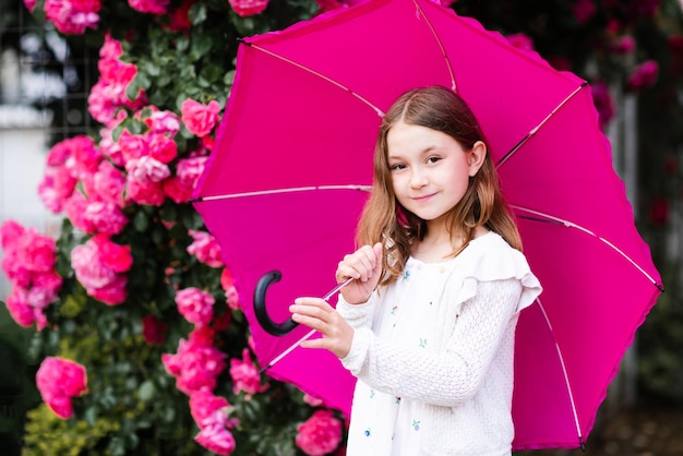 Uśmiechnięta dziewczynka trzyma parasol stojący na ulicy miasta w parku nad kwiatami róż na zewnątrz z bliska