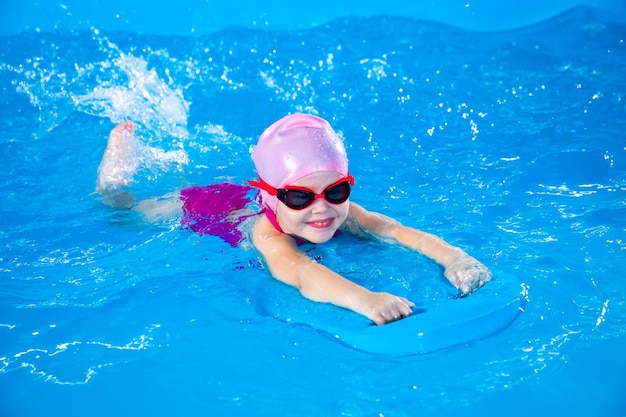 Uśmiechnięta dziewczynka uczy się pływać w basenie z deską pływacką podczas lekcji pływania