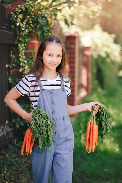 Uśmiechnięta dziewczynka w ogrodzie trzyma dwie kiście świeżych marchewek Pionowo