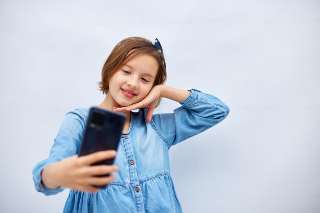 Uśmiechnięta dziewczynka w przypadkowej dżinsowej sukience trzyma smartfona i robi selfie