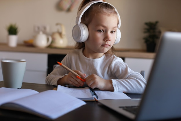 Uśmiechnięta Dziewczynka W Słuchawkach Pismo Ręczne Studiować Online Za Pomocą Laptopa W Domu, Słodkie Szczęśliwe Małe Dziecko W Słuchawkach Wziąć Lekcję Internetową Lub Klasę Na Komputerze.