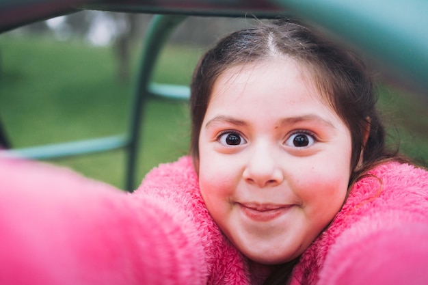Uśmiechnięta dziewczynka z śmieszną buzią robi selfie na placu zabaw w parku.