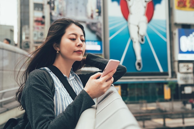 Uśmiechnięta elegancka azjatycka kobieta opierając się na poręczy i wysyłając SMS-y telefon komórkowy online na czacie z uciekinierem znak tablica reklamowa na tle. młoda dziewczyna z telefonem komórkowym na moście dotonbori.