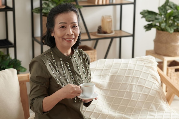Uśmiechnięta elegancka starsza kobieta z filiżanką herbaty siedząca na kanapie w salonie