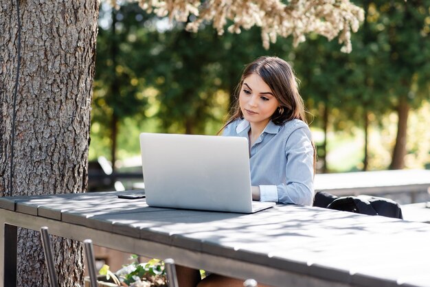 Uśmiechnięta europejska bizneswoman pracuje na laptopie na świeżym powietrzu Piękna młoda dziewczyna siedzi przy stole w słonecznym parku Współczesny styl życia kobiety Koncepcja pracy zdalnej i niezależnej