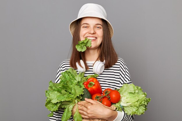 Uśmiechnięta głodna kaukaska kobieta ubrana w pasiastą koszulę i panamę trzymająca organiczne warzywa stojące odizolowane na szarym tle utrzymuje dietę jedząc zieloną sałatę