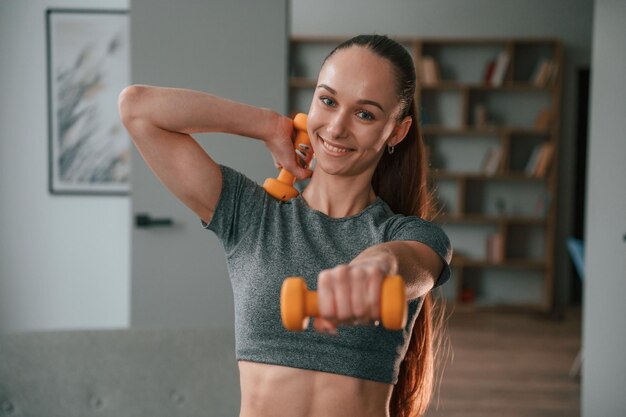 Uśmiechnięta I Trzymająca Hantle Młoda Kobieta W Ubraniach Jogowych Robiąca Fitness W Pomieszczeniu