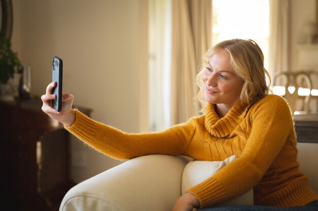 Uśmiechnięta kaukaska kobieta siedząca na kanapie, relaksująca się w salonie, robiąca selfie ze smartfona, spędzająca wolny czas w domu z technologią.