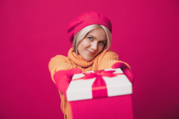 Uśmiechnięta kaukaska kobieta w beretze wyciąga pudełko z prezentami do kamery odizolowane na różowym tle...