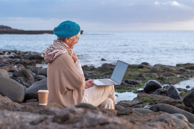 Uśmiechnięta kaukaska starsza kobieta w pracy zdalnej siedzi na plaży za pomocą laptopa Starsza dama z siwymi włosami, ciesząc się morzem, wakacje wideo, rozmowy z komputerem