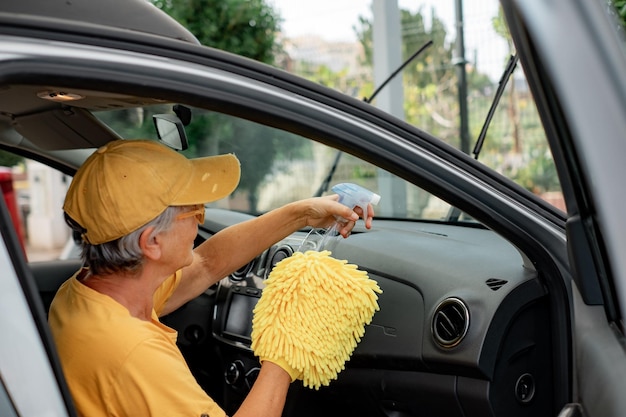 Uśmiechnięta kaukaska starsza kobieta w żółtej koszulce i kapeluszu czyści samochód w samoobsługowej myjni dbając o szczegóły Samoobsługowa myjnia samochodowa