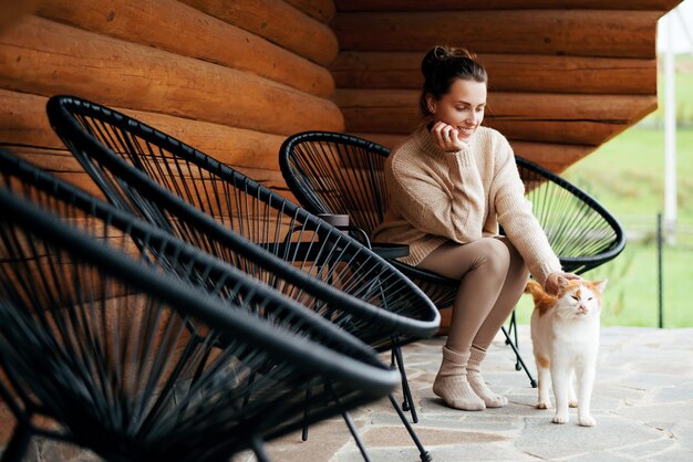 Uśmiechnięta kaukazyjska kobieta pieści dość puszystego kota, siedząc na zewnątrz w pobliżu drewnianego domu Szczęśliwa młoda młoda dama spędza wolny czas na przytulnym tarasie ze swoim uroczym zwierzęciem domowym