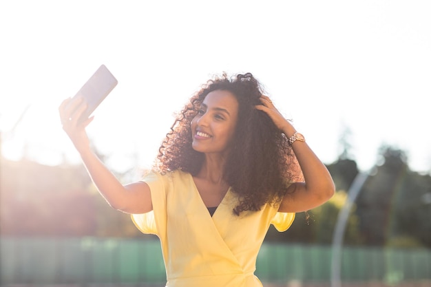 Uśmiechnięta kobieta Afro z kręconymi włosami robiąca selfie podczas zachodu słońca zawiera miejsce na kopię i tekst