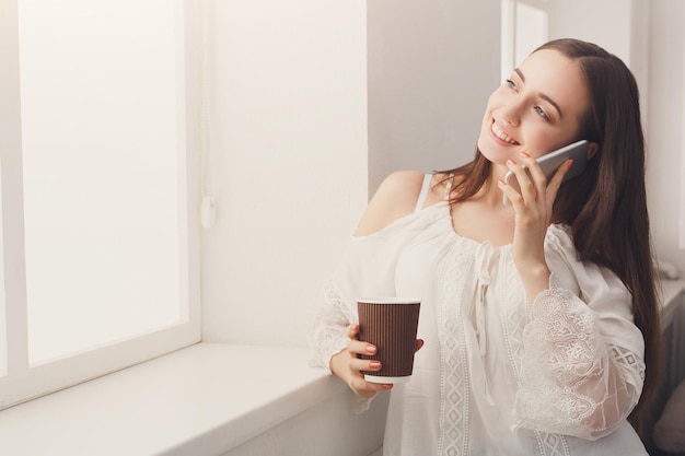 Uśmiechnięta kobieta dzwoniąca na telefon komórkowy, stojąca w pobliżu okna w domu, pijąca kawę, kopiująca przestrzeń